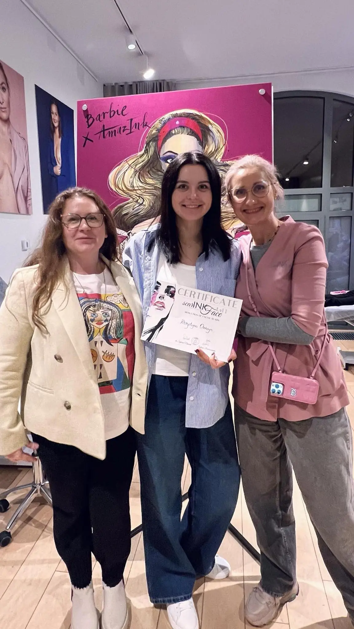 Trzy kobiety stoją razem w pomieszczeniu, a kobieta w środku trzyma zaświadczenie o niedawnym szkoleniu scarink. W tle plakat przedstawiający blondynkę z bliznami. Wszyscy mają uśmiechnięte twarze.