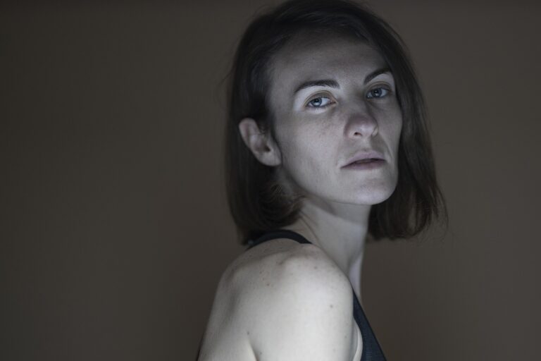 Osoba o krótkich brązowych włosach i neutralnym wyrazie twarzy stoi bokiem, ubrana w ciemny podkoszulek, na prostym brązowym tle.