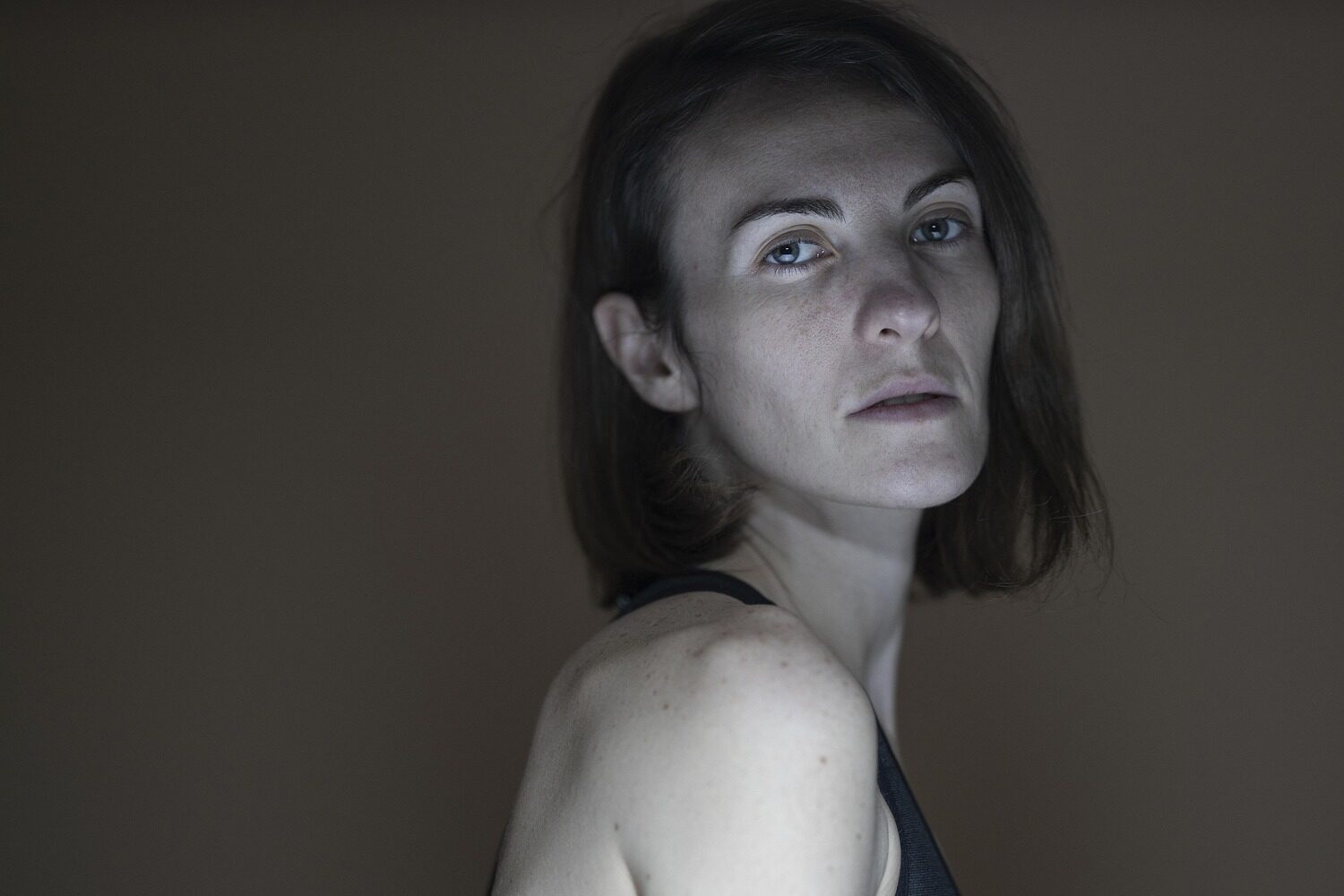 Osoba o krótkich brązowych włosach i neutralnym wyrazie twarzy stoi bokiem, ubrana w ciemny podkoszulek, na prostym brązowym tle.